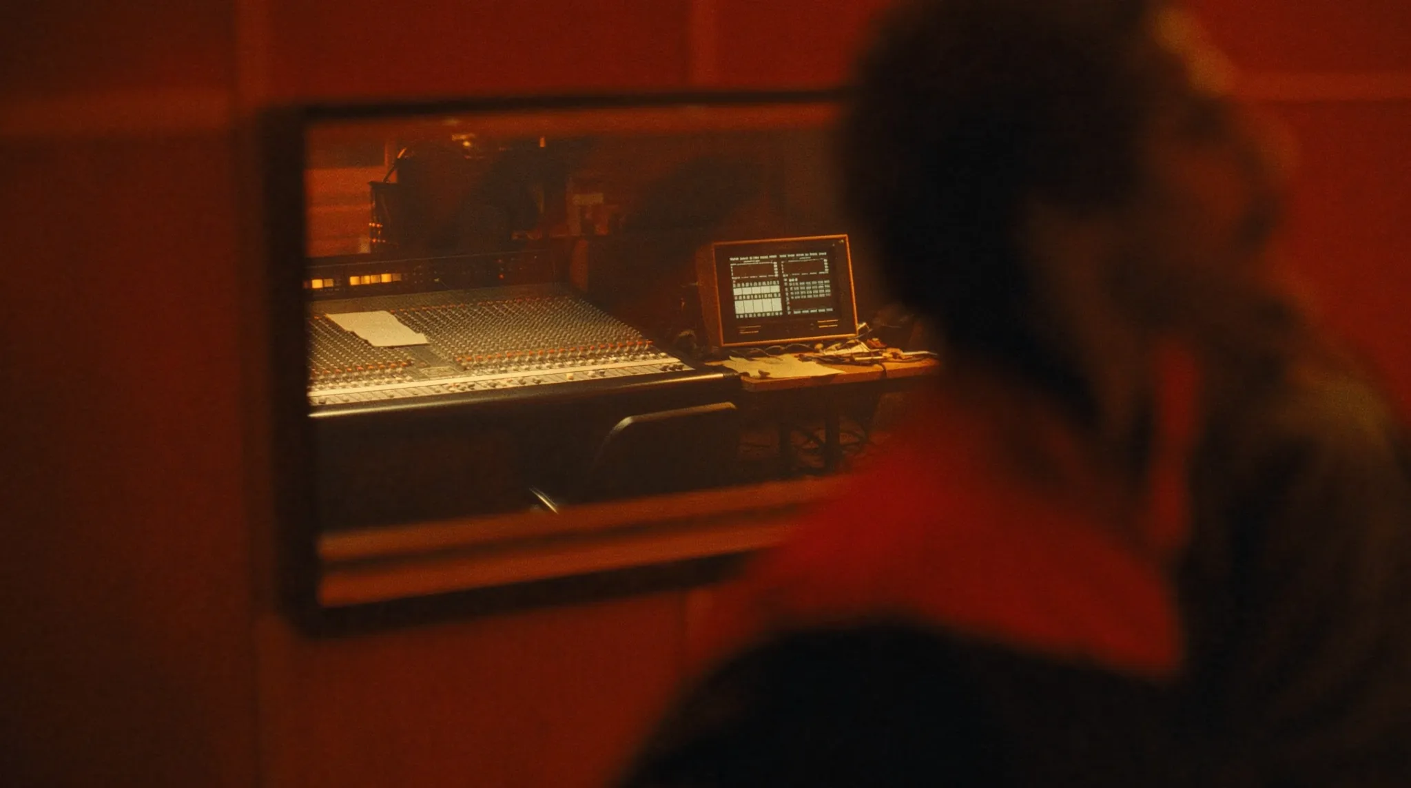 A recording studio control room in warm light: a large mixing console and a monitor seen through the glass.